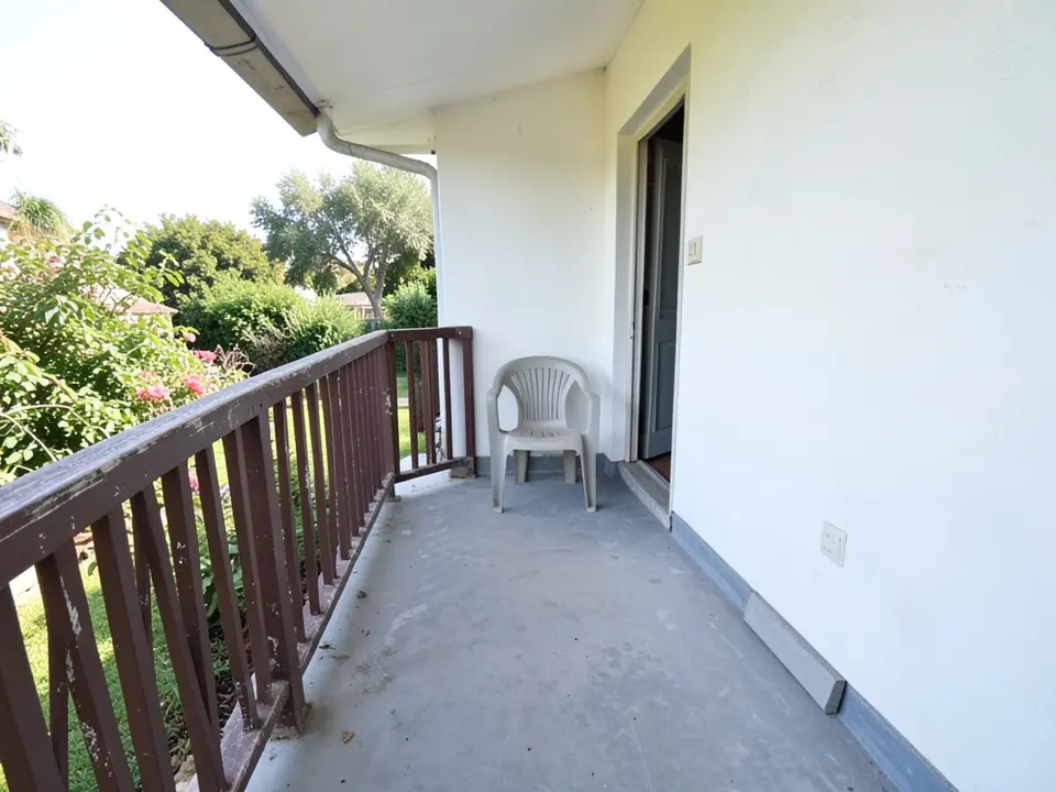 A house balcony with an old brown painted wooden railing with peeling paint, a grey concrete floor, a white wall with a door, a white plastic chair and a garden with a flowering shrub beyond