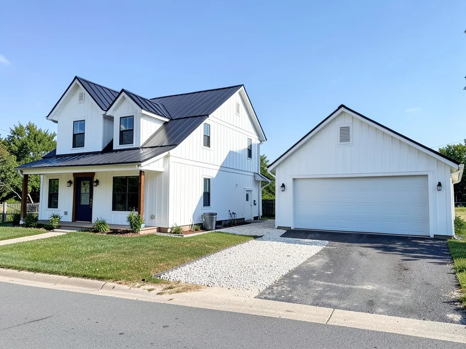 A white two-storey modern farmhouse with a black metal roof on the left and a separate white garage on the right, with a gravel strip between them