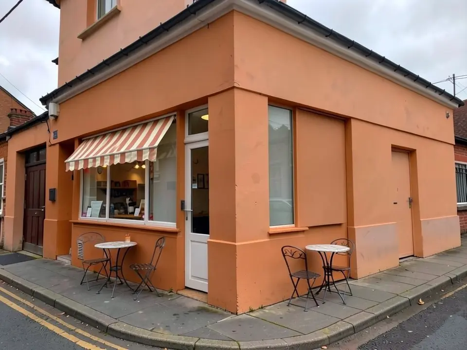 The same corner building painted terracotta orange, with a striped cream and terracotta awning over the window and two small round bistro tables with chairs on the pavement, and the same maroon door, black gutter and street