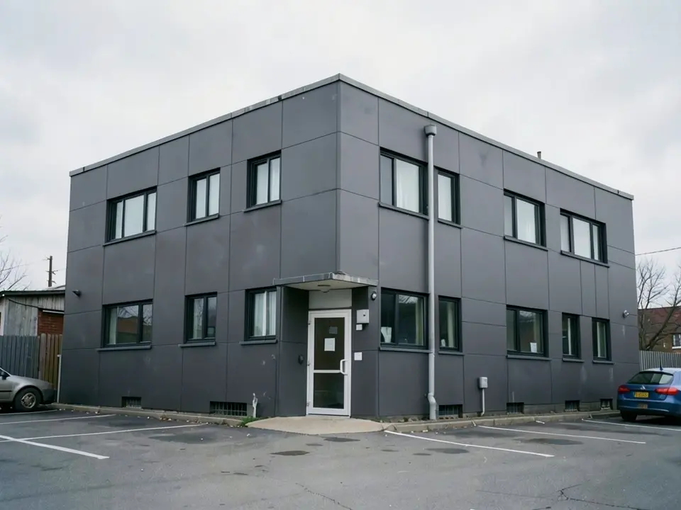 The same office building with dark grey cladding panels with visible joints instead of brick, black window frames, and the same white entrance door and canopy, drainpipe, car park and cloudy sky