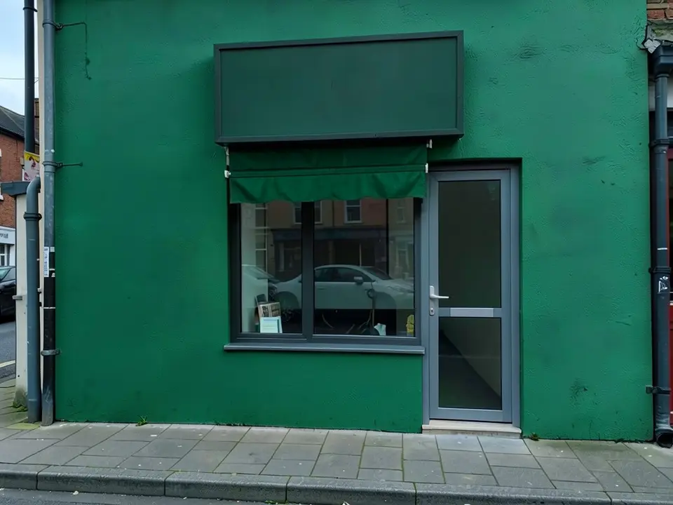 The same shop front with a deep green wall, a dark green blank signboard, a green fabric awning with a scalloped edge over the top of the window, and the same grey door, drainpipes and pavement