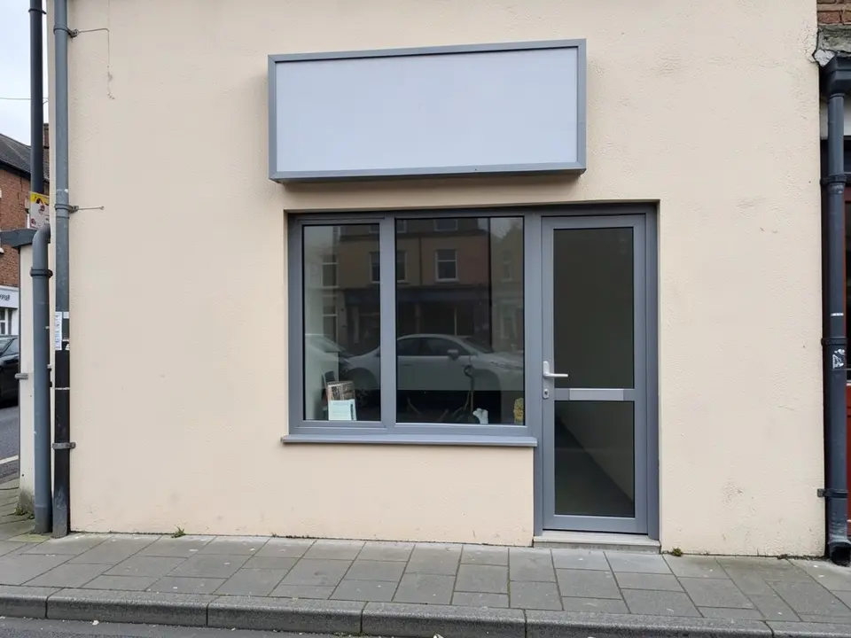 A small shop front on a street corner with a cream rendered wall, a blank pale grey signboard, a large window with a grey frame, a grey glass door, grey drainpipes and a paved pavement