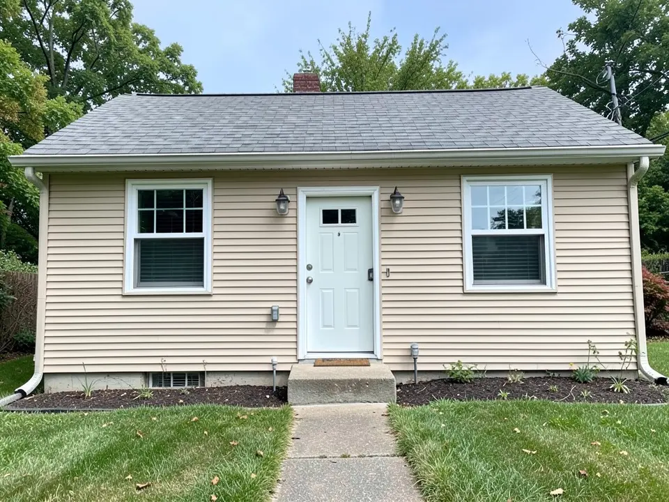 Front of a small single-storey house with beige horizontal siding, a low grey shingle roof with a brick chimney, two white-framed windows, a white front door with two wall lanterns, a small concrete step, mulched beds, a lawn and a short path