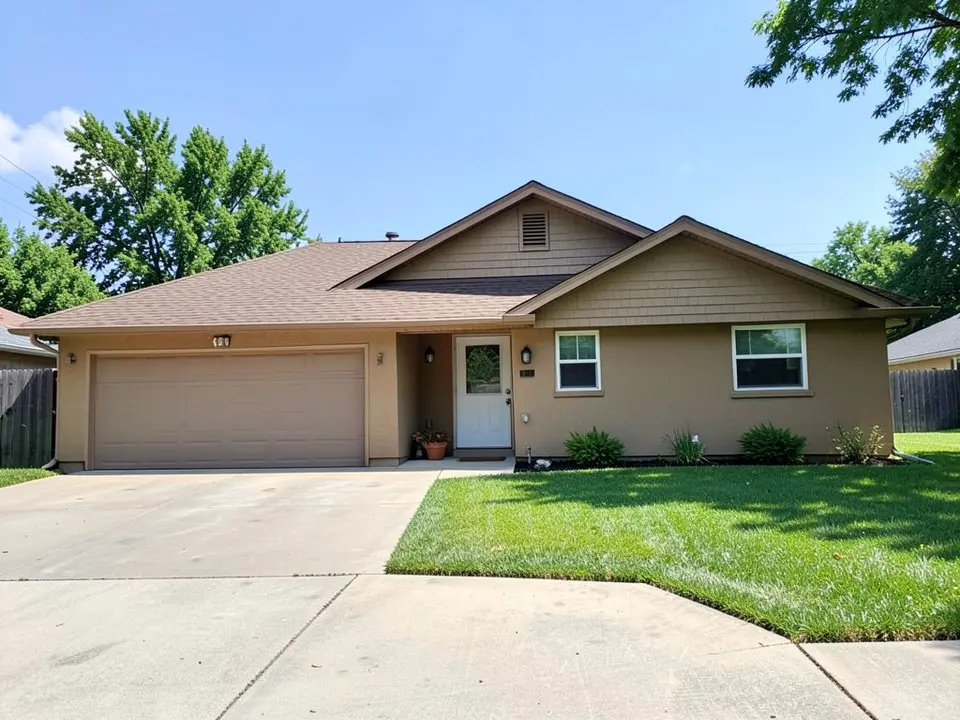 Front of a single-storey tan stucco ranch house with a low brown shingle gable roof, a double garage door on the left, a white front door, two small windows, a lawn and a wide concrete driveway