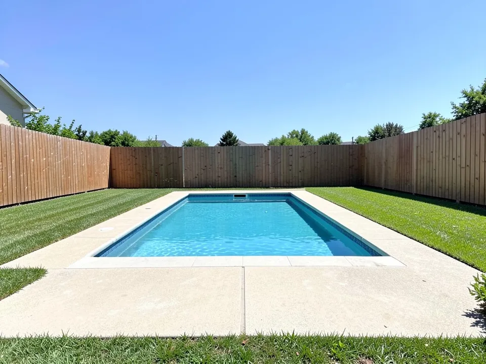 A backyard with a rectangular pool, a concrete deck, lawn on both sides and a tall wood fence at the far end, with nothing built