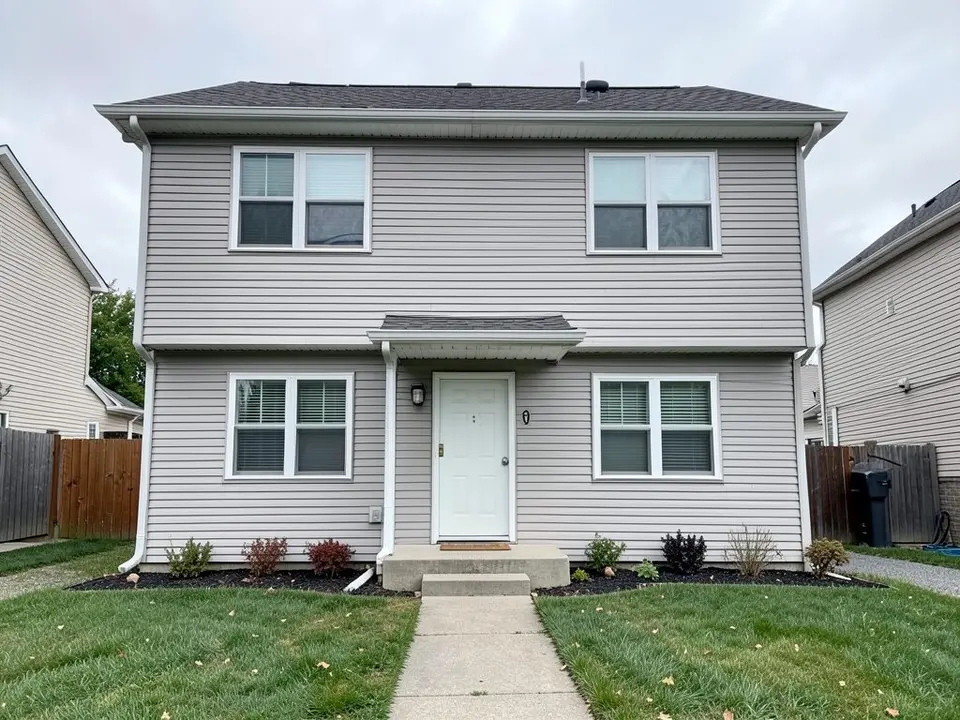 Front of a two-storey house with light grey siding, a white front door with a small shingled hood above it, a concrete step and a path, two windows upstairs and two downstairs, small shrubs along the wall and a lawn