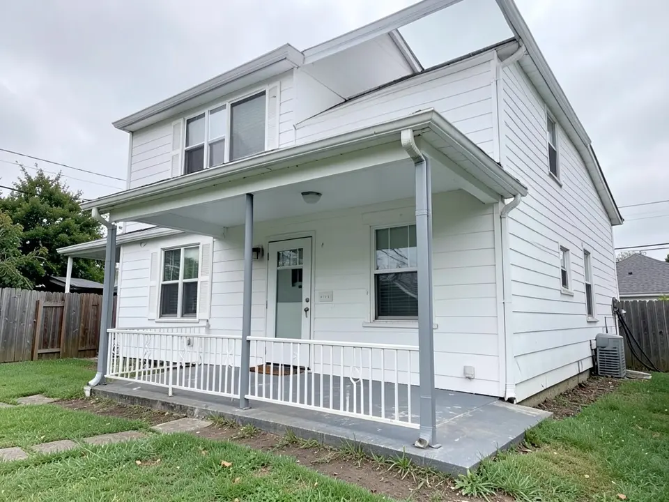 Front of a white clapboard two-storey house with a covered porch held up by three thin grey posts, a white metal railing with a scroll pattern, a grey painted concrete floor, a white ceiling and a pale front door, with a lawn and stepping stones