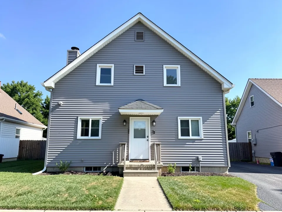 A grey vinyl-sided two-storey house with a steep front gable, a small hood over the door, white windows, a lawn, a path and neighbouring houses