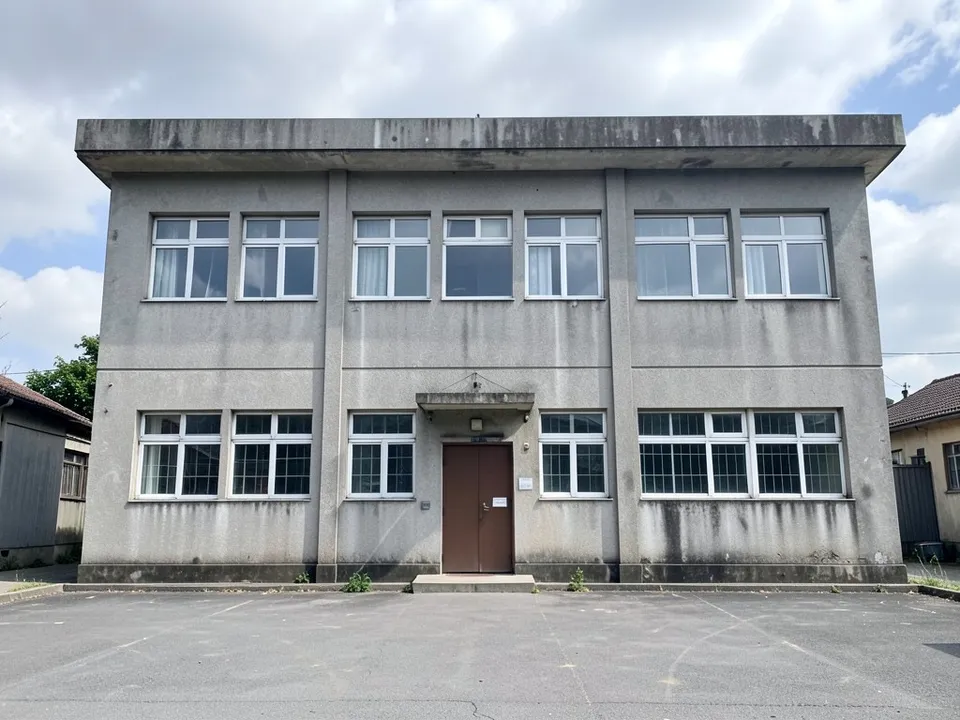 An old stained concrete two-storey school block with rows of small windows, a flat overhanging roof, a small door with a canopy and a tarmac yard