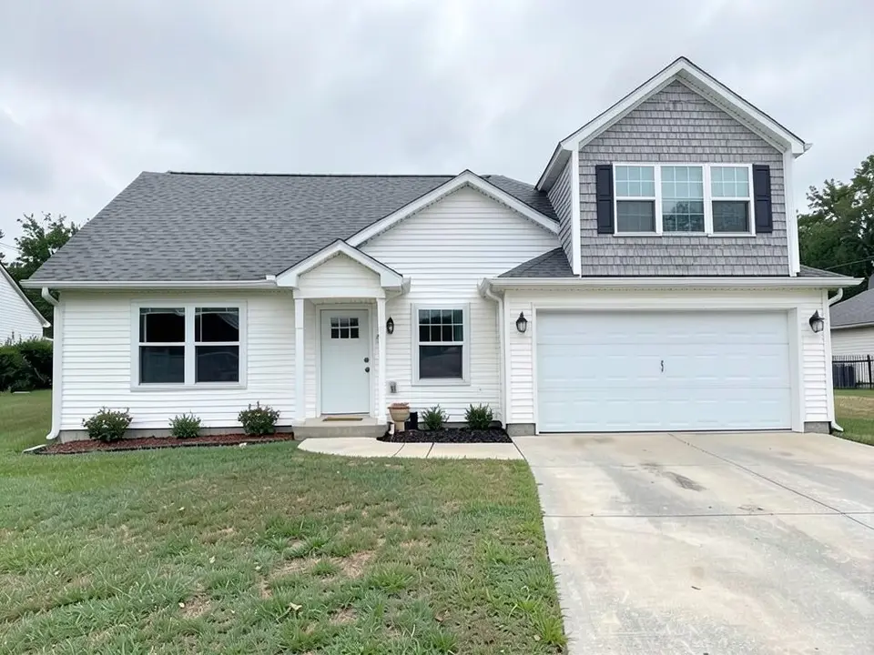 The same house with a grey shingle-clad upper floor with a gable roof over the garage wing, three windows in a row with black shutters on each side, and a short lower roof across the garage front, with the same garage door, front door, windows, driveway and lawn