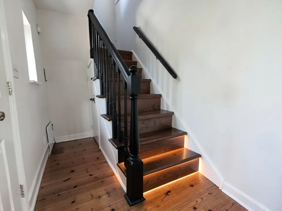 The same staircase with dark walnut-coloured treads and risers, black balusters, a black newel post and black handrails, and a warm glowing light strip under the lowest steps, over the same pine floor