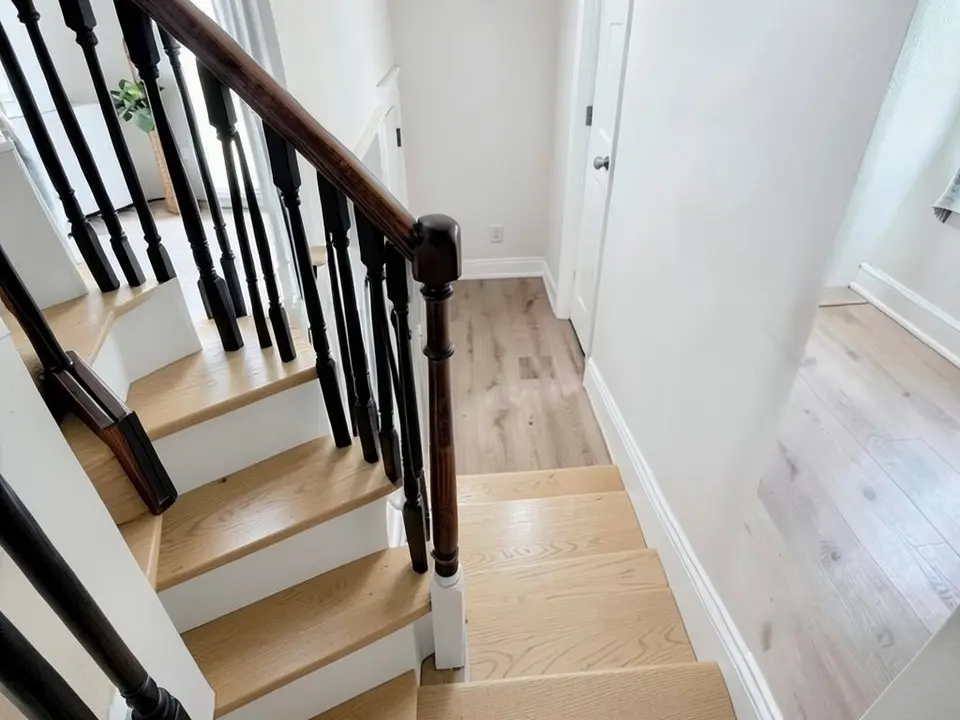 The same staircase with the carpet gone, light oak-coloured wooden treads with white risers, slim black balusters under the same dark brown handrail, and the same white walls and floor