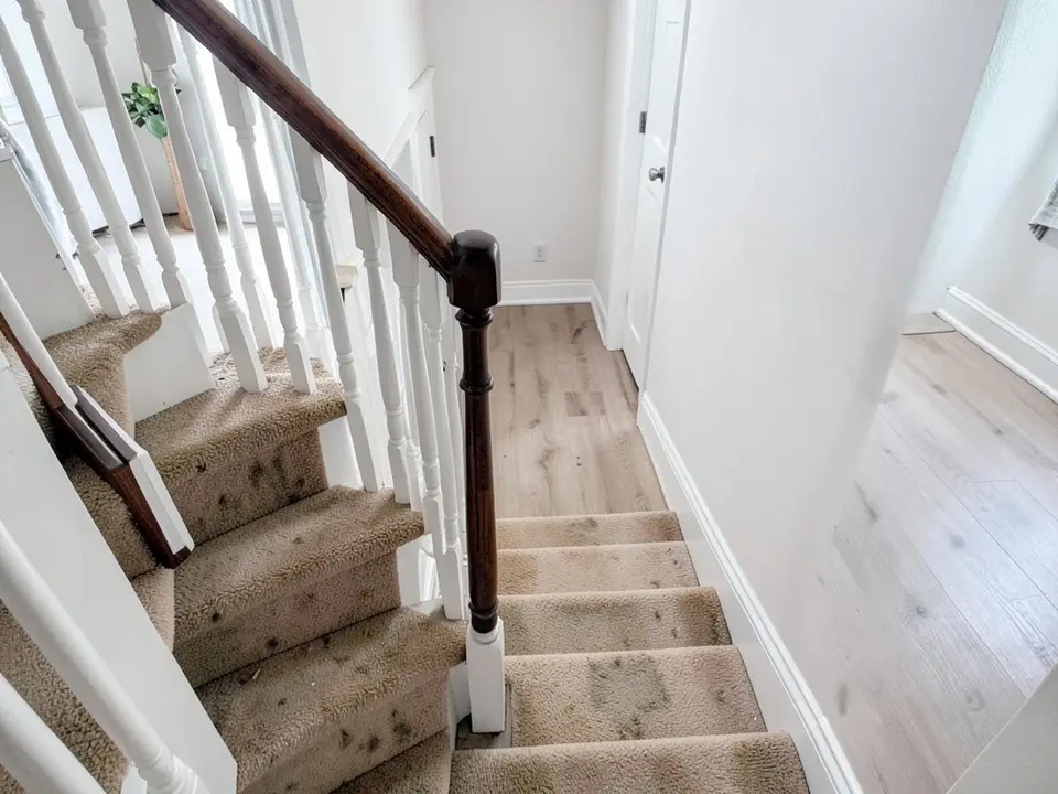 A staircase seen from above with stained beige carpet on the steps, white spindles, a dark brown handrail and newel post, white walls and a light wood-look floor at the bottom