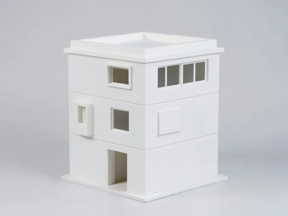 A plain white clay model of a three-storey corner house with a flat roof terrace behind a parapet, a recessed entrance and a small projecting box window on the middle floor