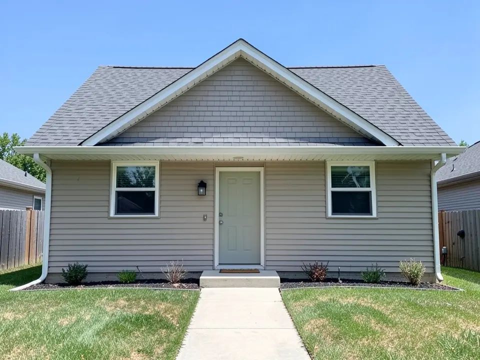 Front of a house with beige horizontal siding, a grey shingle gable roof with short eaves, a pale front door, a lawn and a concrete path
