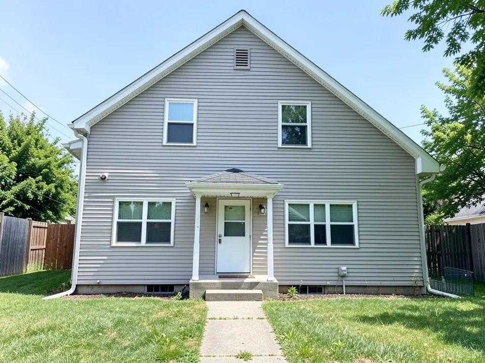 Front of a plain two-storey house with pale grey horizontal siding, a steep gable with a vent, a small concrete stoop with a little roof on two thin white posts, white-framed windows, a lawn and a short path