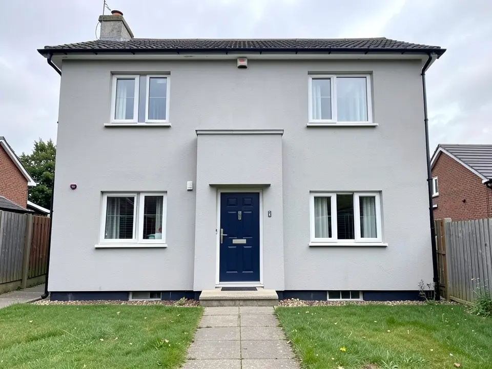Front of a two-storey house with light grey rendered walls, a dark blue front door inside a slightly projecting rendered surround with a small ledge above it, four white-framed windows, a dark tiled roof with a chimney, wooden fences on both sides, a path and two small lawns