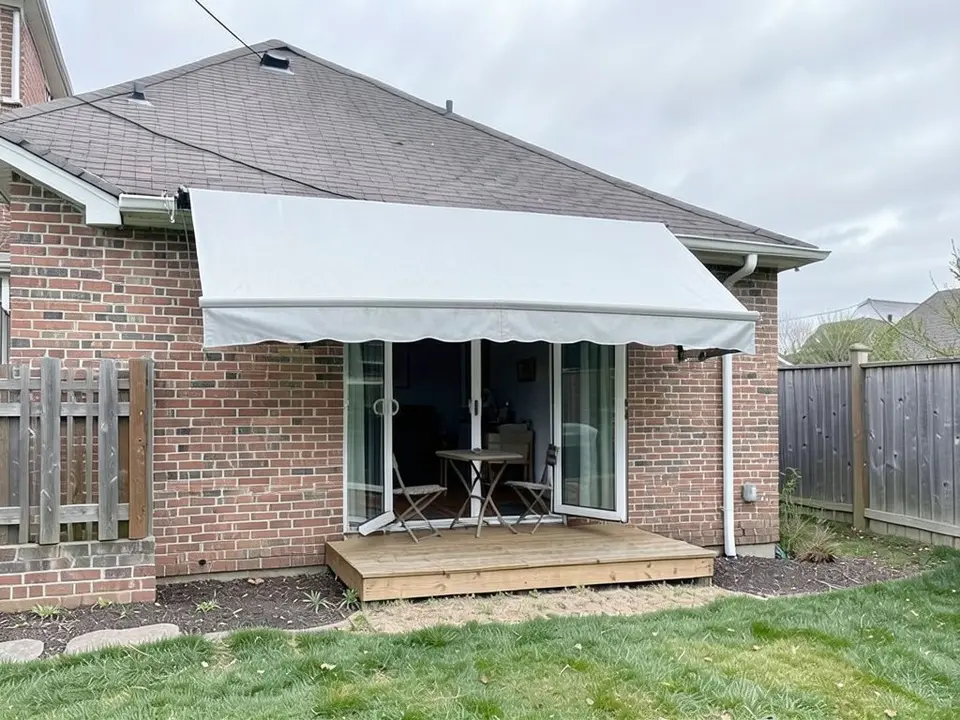 The same house with a large light grey retractable-style awning sloping down from under the roof edge over the patio doors and the deck, with a front bar and a scalloped valance, and the doors shown closed behind it, with the same deck, table, chairs, fences and lawn