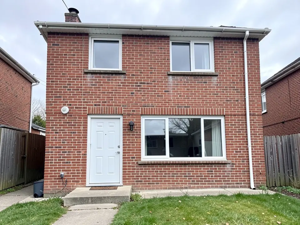 Back of a two-storey red brick house with a white door and a concrete step, a large white-framed ground-floor window, two white-framed windows upstairs, a white downpipe on the right, wooden fences on both sides and a small lawn