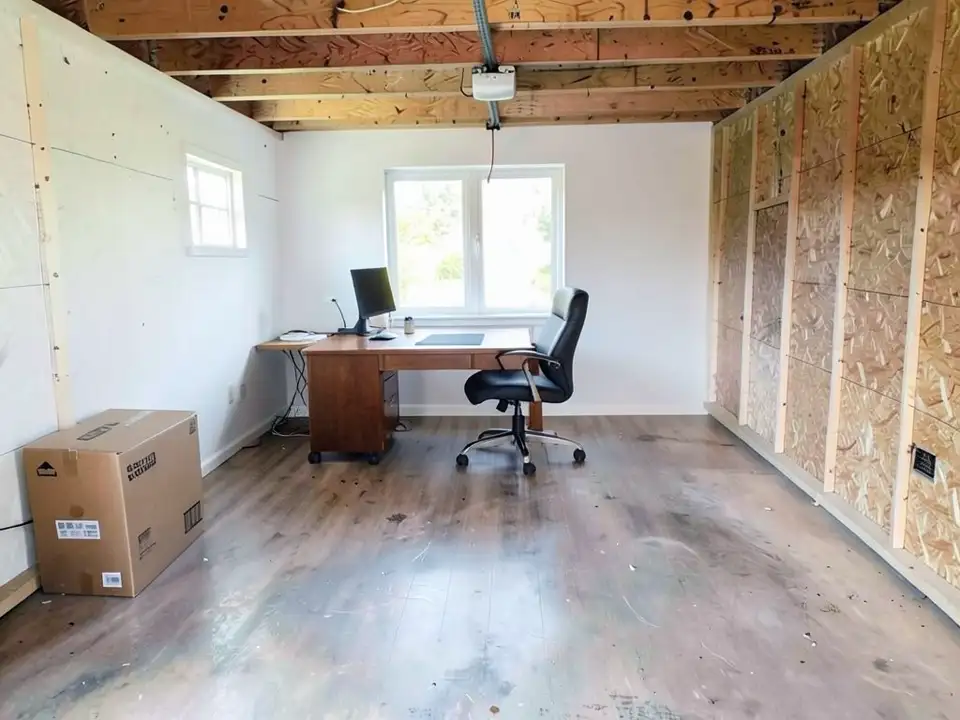 The same garage interior with white-painted walls on the left and back, a wood-look floor, a wooden desk and black chair under a new window at the far end, and the right wall and ceiling still bare