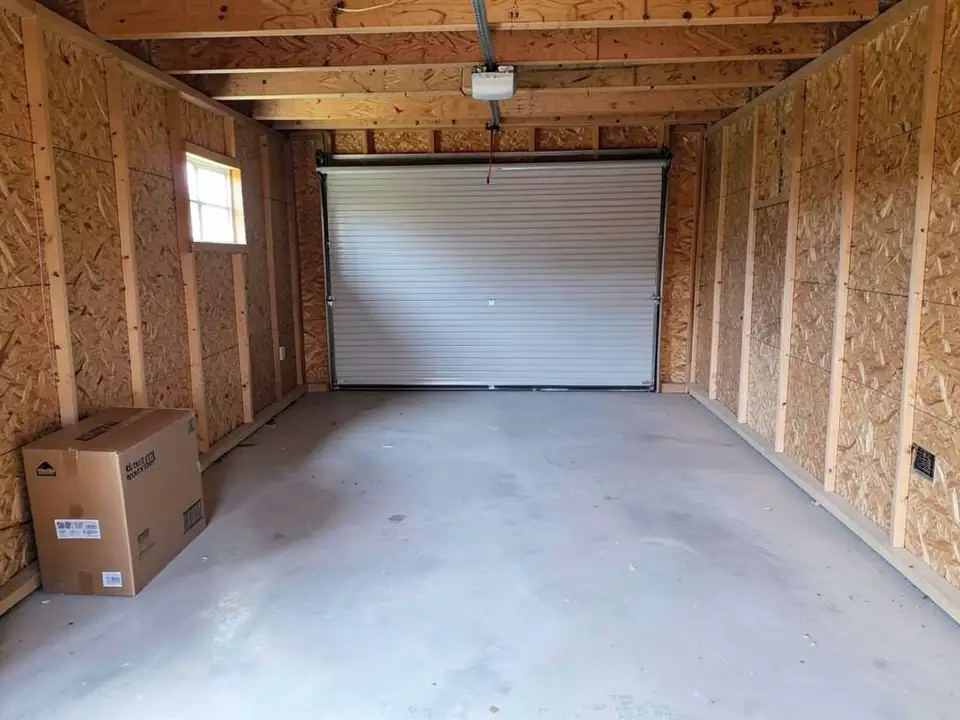 Inside a bare single-car garage with unfinished wood-panel walls, a concrete floor, exposed ceiling joists, a closed roll-up door at the far end, a small window on the left and a cardboard box