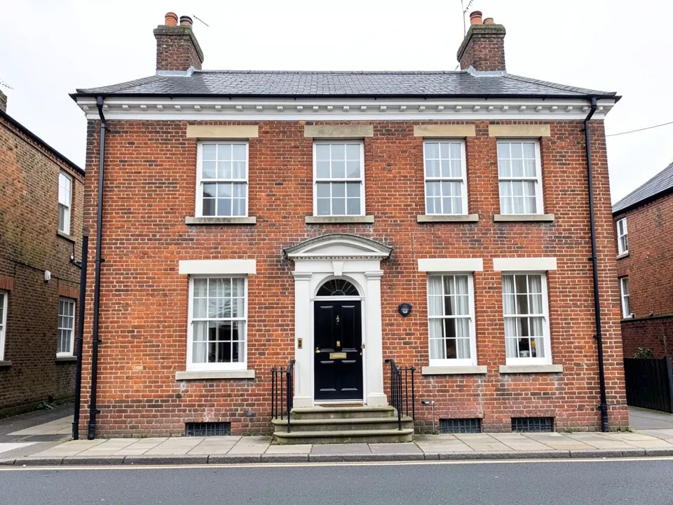 Street photo of a two-storey red brick Georgian house with four sash windows upstairs, one window left of a black front door and two to the right, a white door surround with a fanlight, stone steps with black railings, two chimneys with pots, a slate roof and brick neighbours on both sides