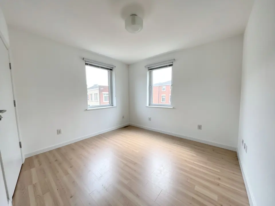 Wide-angle photo-style view of an empty apartment room with white walls, a pale laminate floor, two windows with blinds looking out at brick buildings, a white ceiling light and a door at the left edge